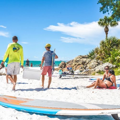 People are relaxing on a sunny beach with surfboards, greenery, and a clear blue sky in the background.