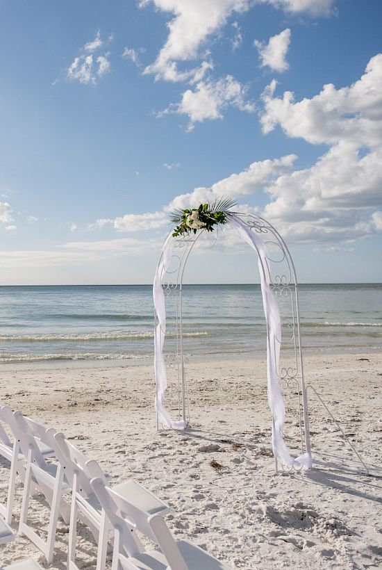 A beach wedding setup with white chairs and an arch facing the sea, under a partly cloudy sky with a boat in the distance.