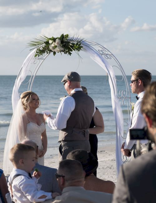 A beach wedding ceremony with a couple under an arch, surrounded by guests and wedding party near the ocean.