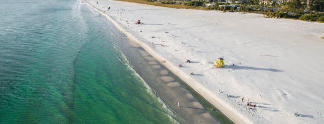 A beach with white sand, green water, scattered people, and a lifeguard station. Buildings and trees in the background.