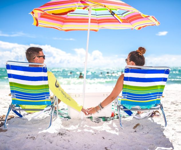 A couple holds hands on a beach, sitting in striped chairs under a colorful umbrella, facing the ocean.