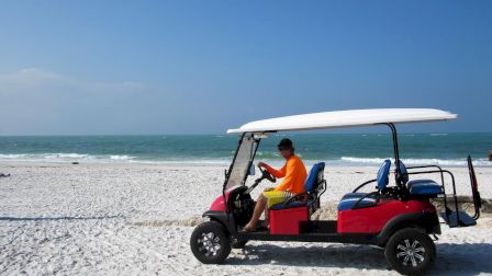 A person in a bright outfit drives a red golf cart with a surfboard on top along a sunny beach with clear blue skies.