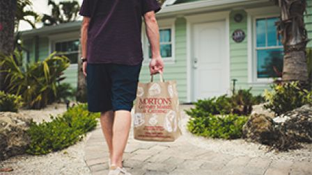 A person is walking towards a house, holding a paper bag with "Morton's Gourmet Market & Catering" on it, surrounded by plants and pathway.