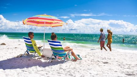 People relax on a beach under a colorful umbrella with chairs facing the ocean, while others walk along the shore in the background.