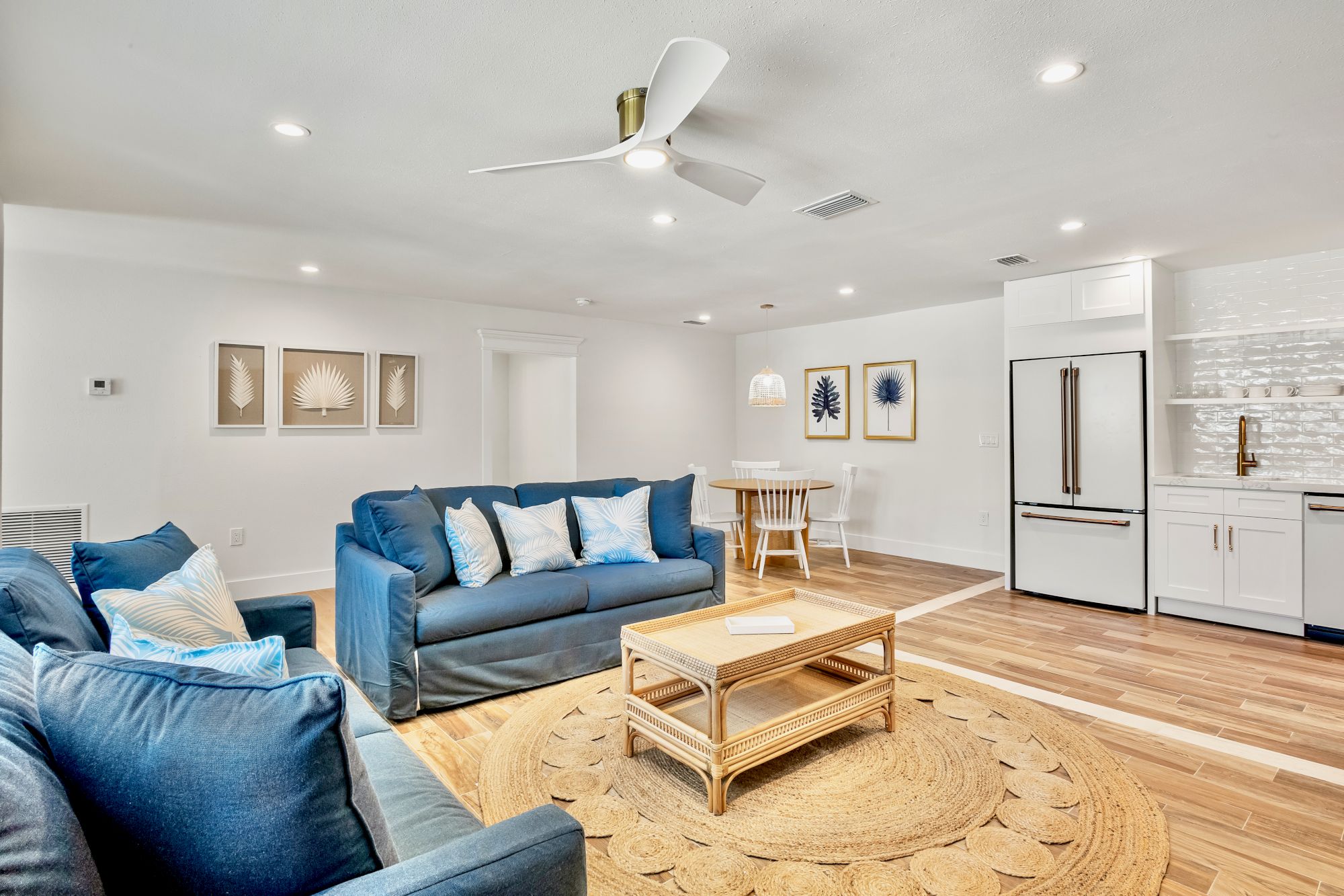 A bright living space with blue couches, a wooden coffee table, wall art, dining area, and white kitchen appliances. Ceiling fan centered.