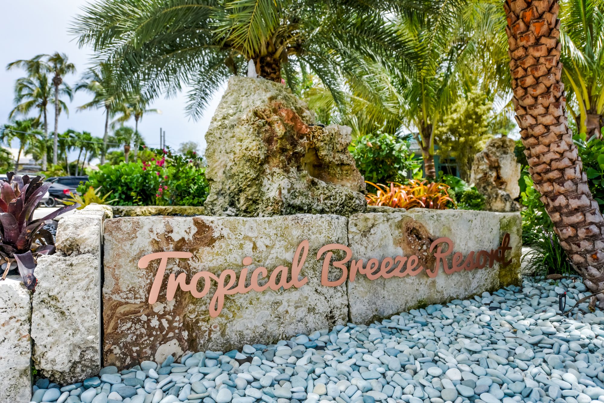 A sign reading "Tropical Breeze Resort" surrounded by palm trees and vegetation, set against rocks and a bed of light stones.