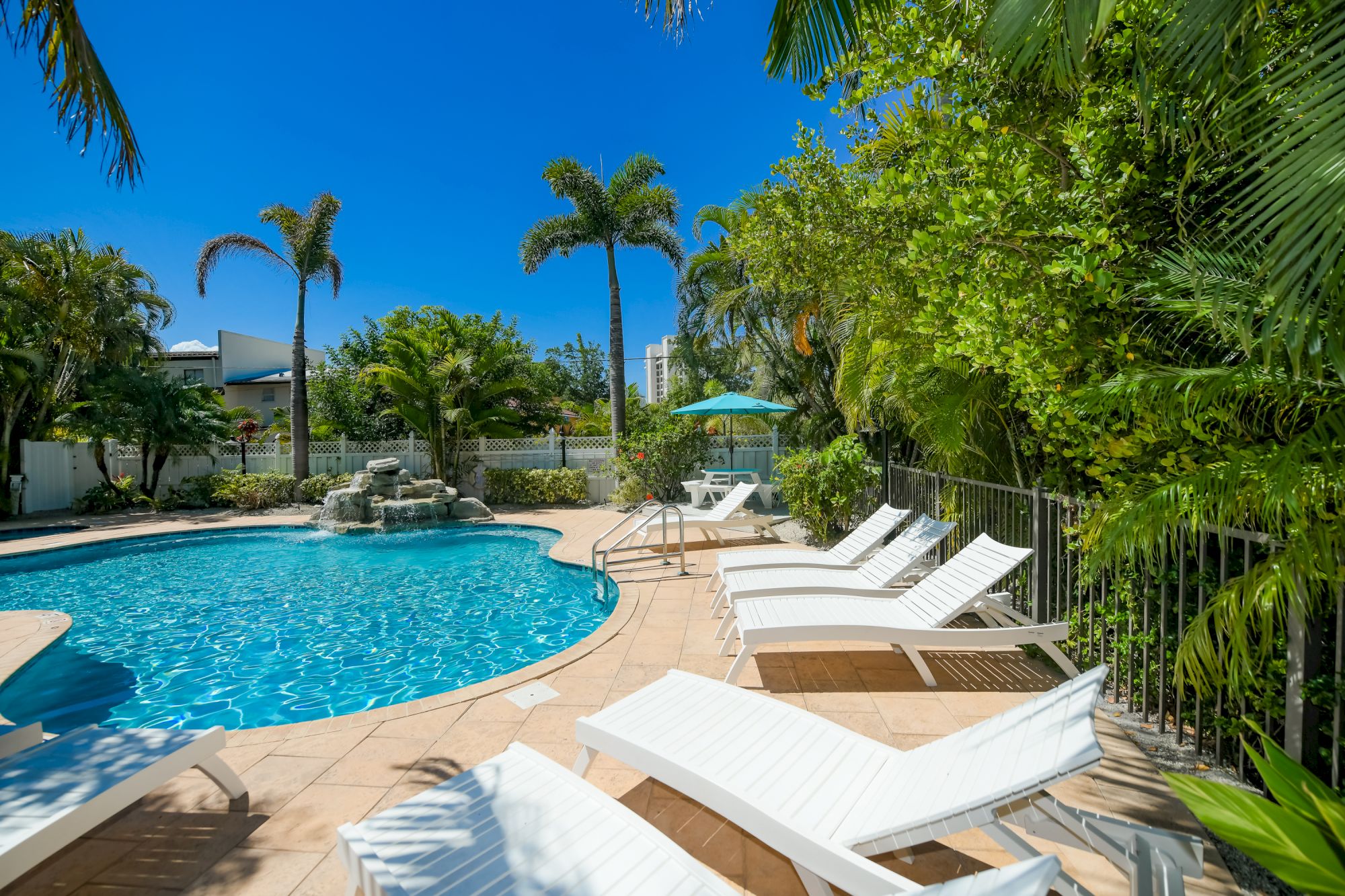 A tranquil pool scene with lounge chairs, tropical plants, and palm trees under a clear blue sky completes the relaxing setting.